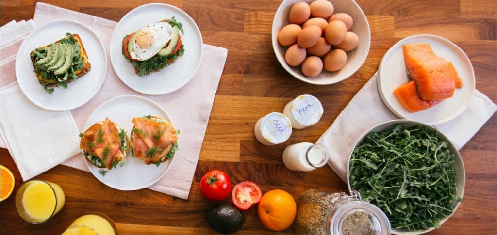 Overhead view of protein-rich foods including eggs, salmon, yogurt, avocado toast, leafy greens, and legumes arranged on a wooden table.