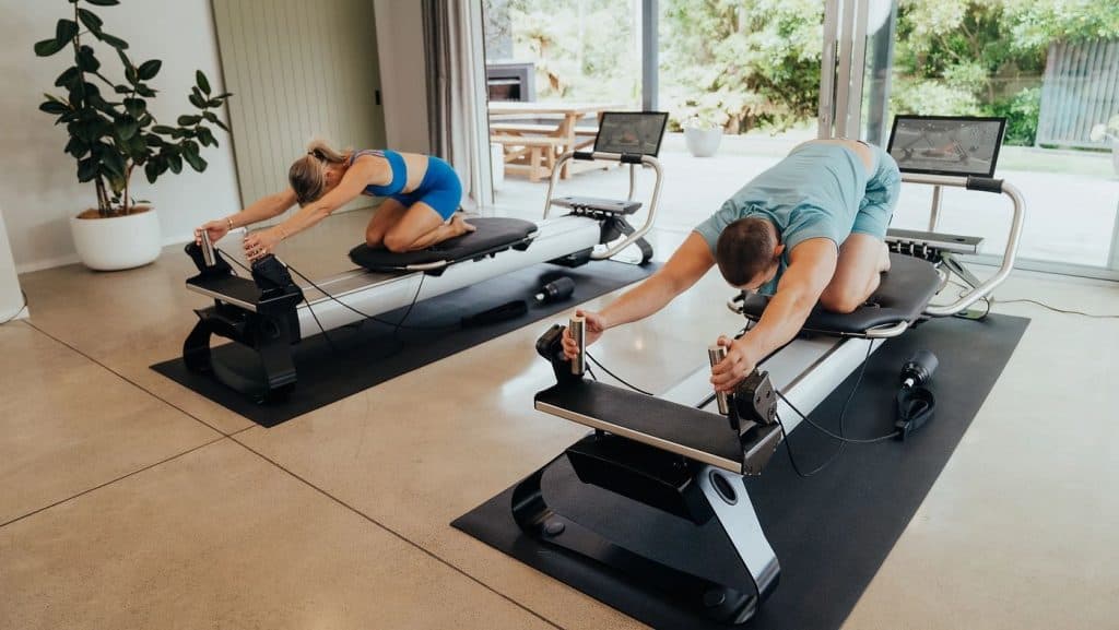 Individuals performing a stretching sequence on connected reformers in a modern home space.