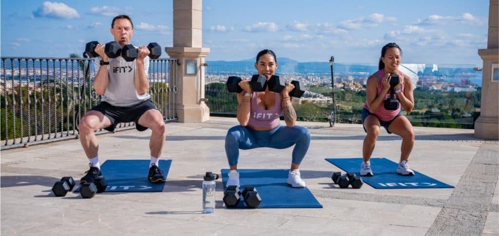 Three iFIT trainers performing dumbbell squats during a guided outdoor strength workout, highlighting structured resistance training.