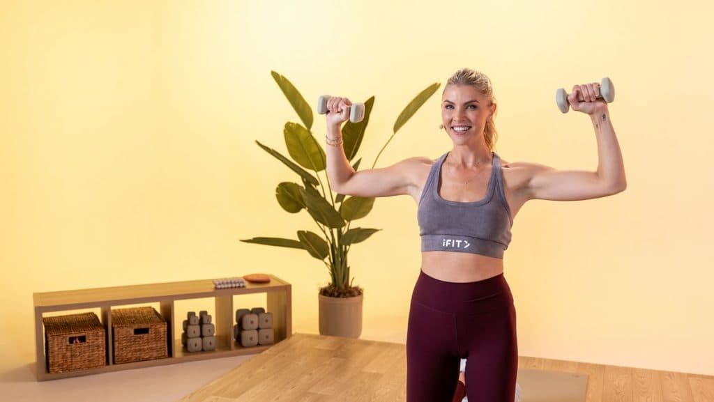 Instructor performing controlled shoulder presses with light dumbbells in a bright studio, highlighting strength-building through intentional movement.