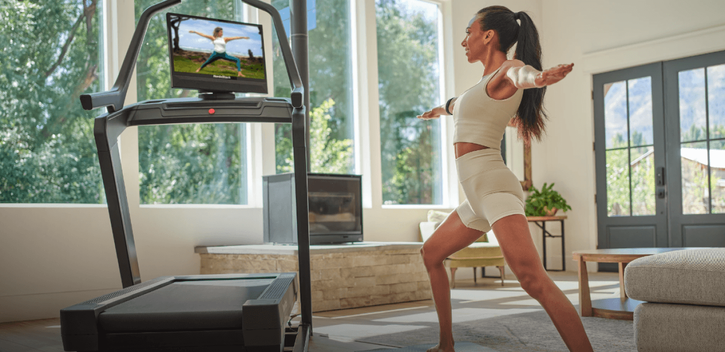 In a beautiful home with large windows, a woman stands next to her treadmill doing yoga, following a trainer on the rotated treadmill screen.