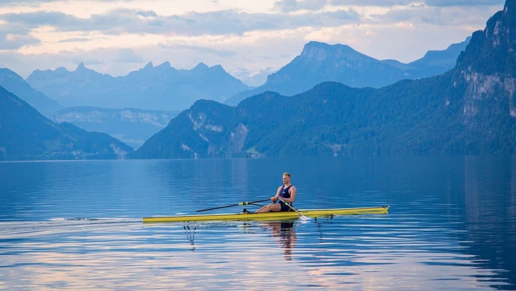 World-class rower training in a single scull on calm open water surrounded by mountain scenery, highlighting endurance and rhythm.